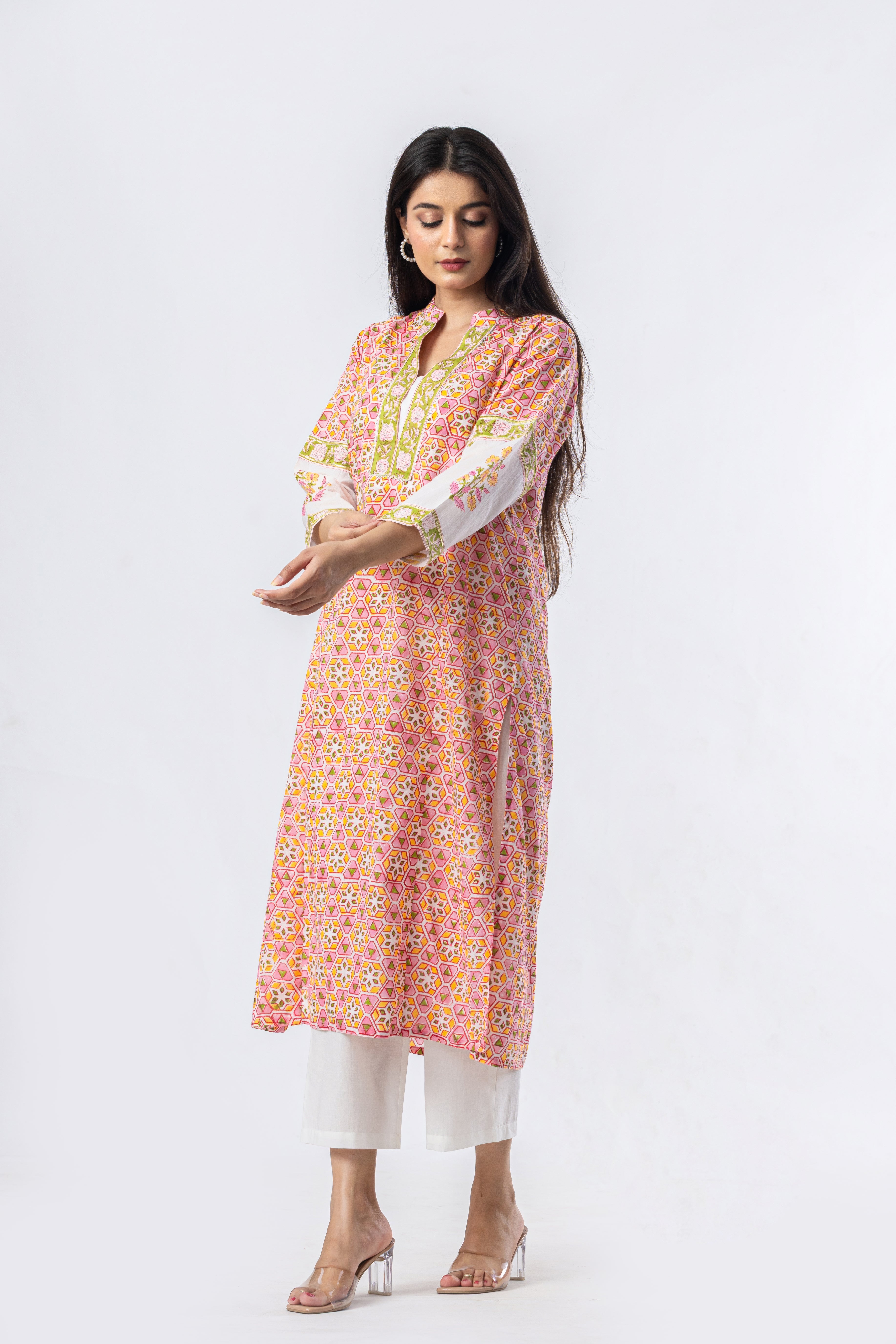 A woman modeling a pink floral printed kurta with yellow and green embroidery on the neckline and cuffs, paired with white pants and clear high-heeled sandals, against a plain white studio background.