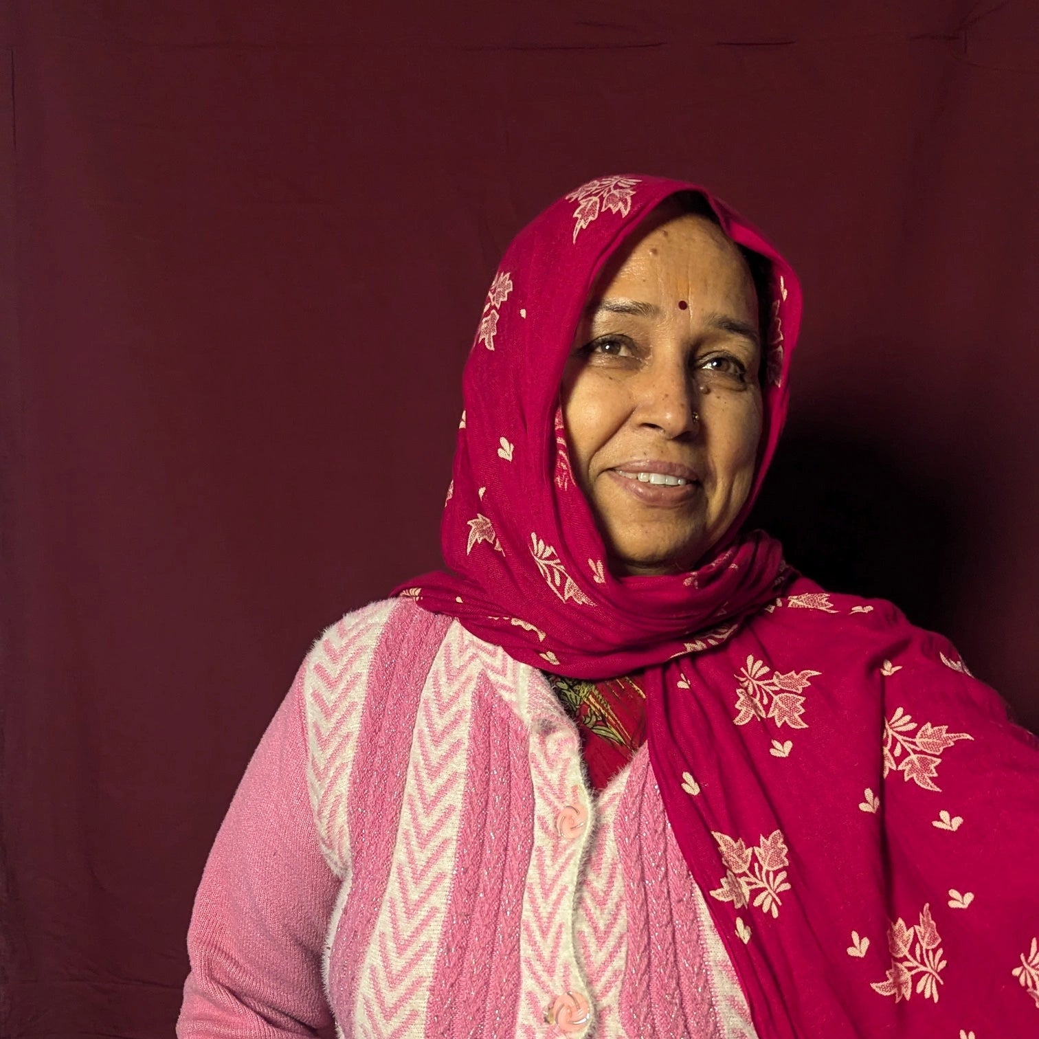 Woman wearing a pink and red traditional outfit with a patterned headscarf against a dark background