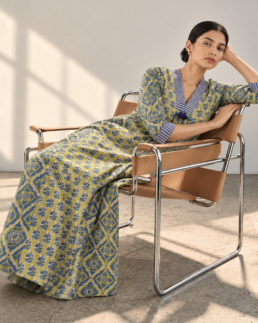 A woman wearing a long-sleeved V-neck maxi dress with a blue and yellow geometric pattern sits on a modern metal-framed chair in a minimalist room with warm sunlight and window shadows.